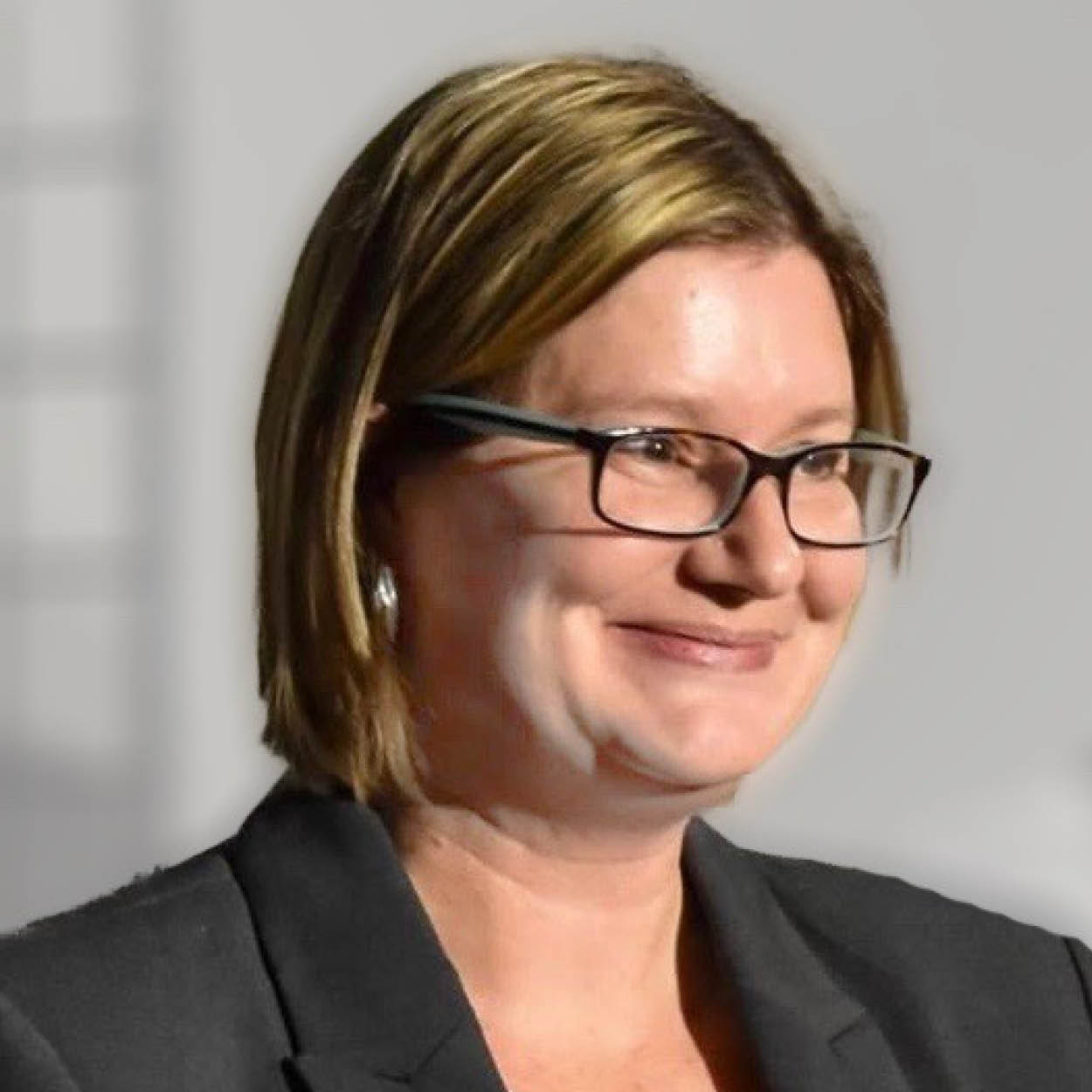 Julia Hendry, a white woman with short blond hair wearing glasses, smiles warmly off camera. She is wearing a black blazer and a Laurier name tag.