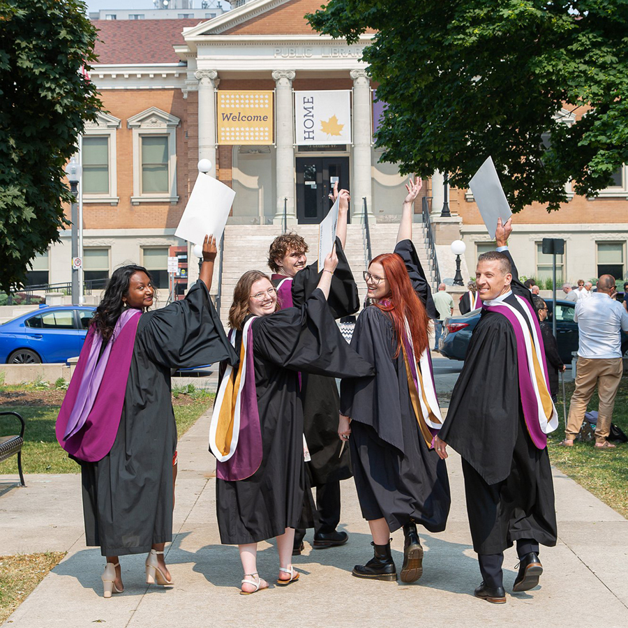Five alumni in Laurier convocation robes triumphantly hold their diplomas in the air and grin at the camera.