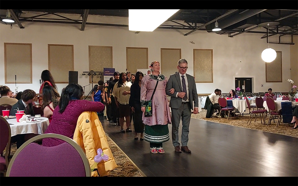 Indigenous dancer Naomi Martin and TD Branch Manager stand together with arms linked. Two lines of dancers stand behind them as they wait for the music to begin.