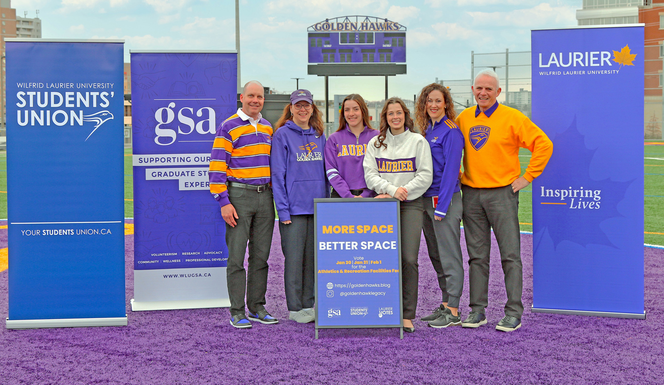 Jason Coolman, Deb McLatchy, Kate McCrae Bristol and Dave McMurray stand on Alumni Field with the presidents of the Laurier Student's Union and Graduate Students' Association.