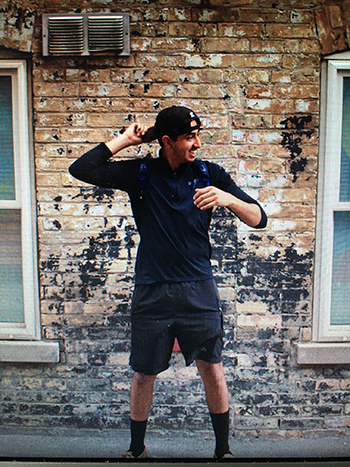 Nathan is pictured outside through a screen door. He is turning his baseball cap backwards on his head and smiling to the right of the camera view