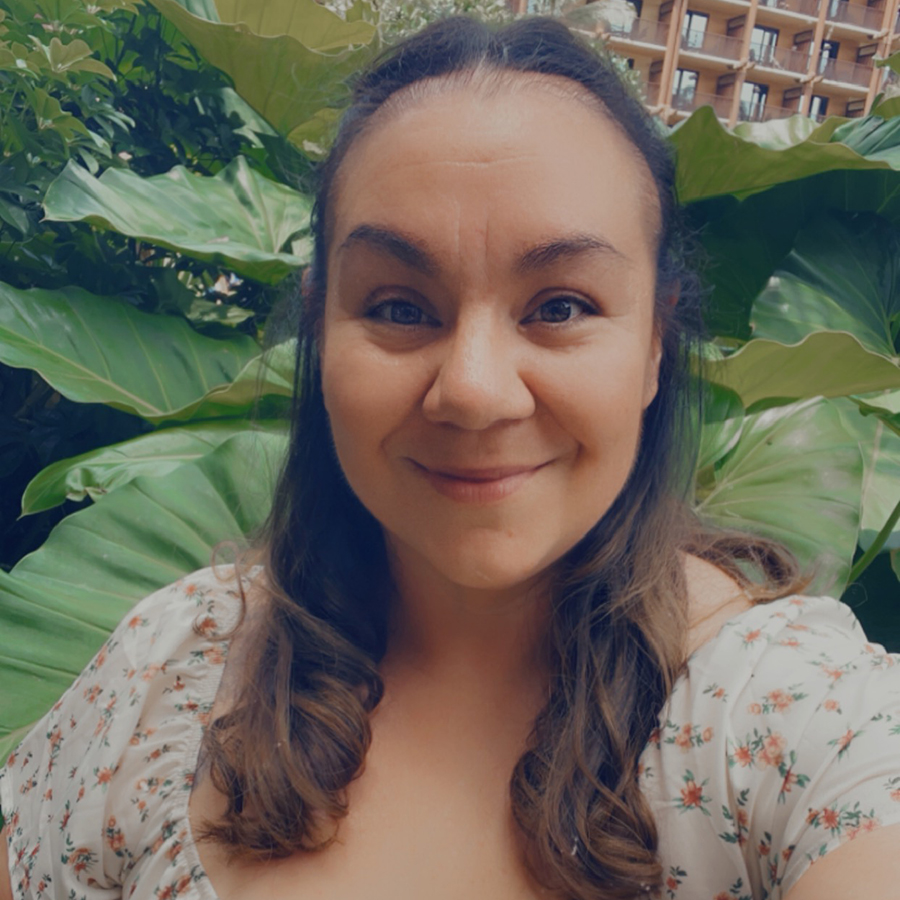 A selfie of Jessica Bomberry, wearing a floral shirt with large plants in the background