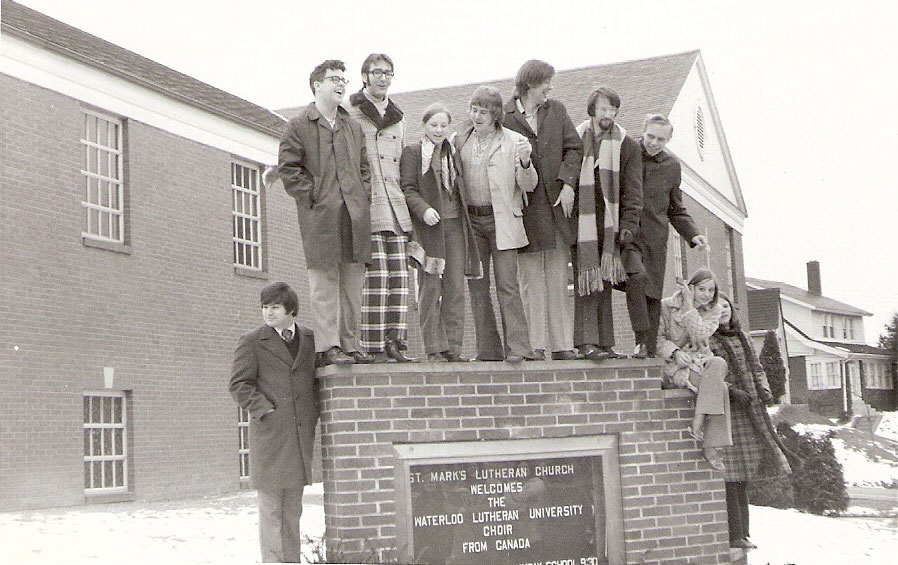 A group of young people standing on and beside a church sign and smiling.