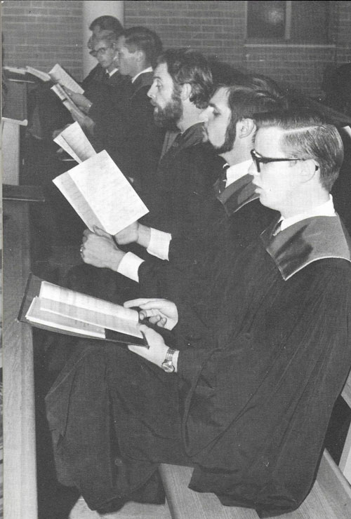 Six young men singing, wearing robes, seated, photo taken from the side.