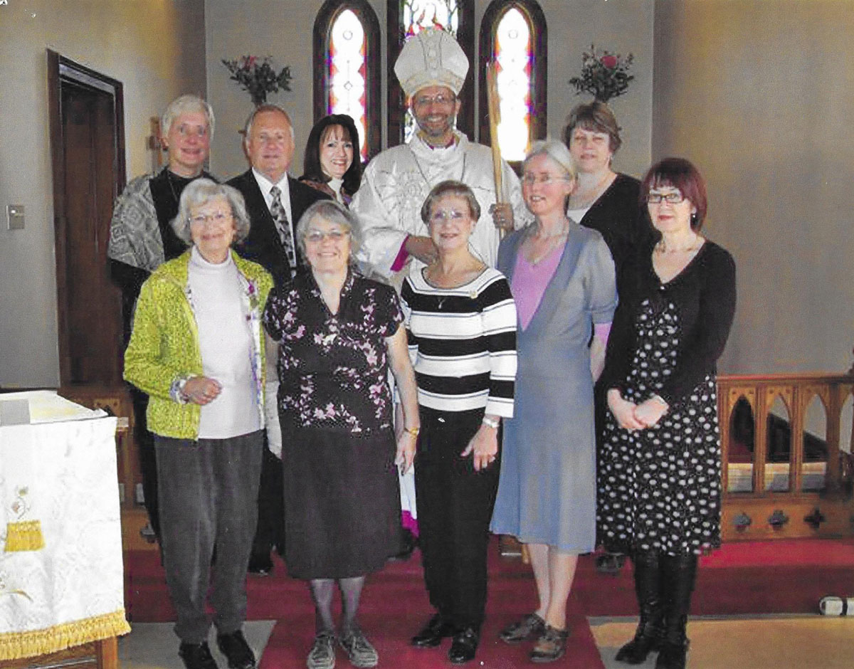 A group of nine people wearing Sunday clothes  posed at the front of a church, with a man in a Bishop's robe and mitre.