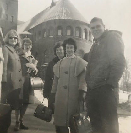 A group of young people in front of a romanesque revival style building, carrying sheet music.
