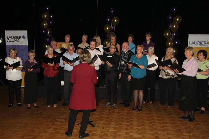 A group of about 25 young and older people singing, with the conductor's back to the audience. The room is dark, has balloons in Laurier colours, a Laurier Alumni sign and a shiny parquet floor.